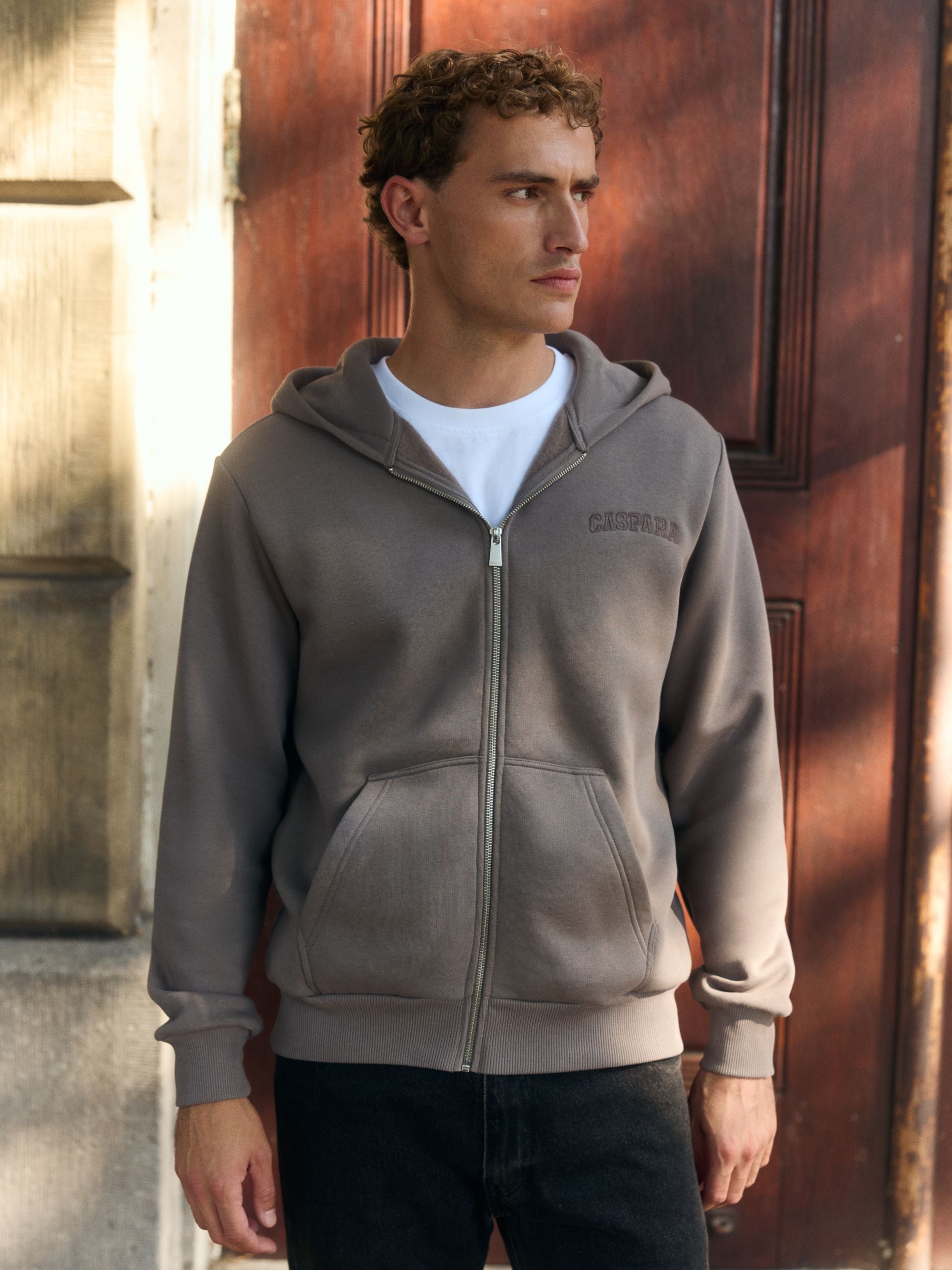 Man wearing a walnut CASPARA logo zip hoodie standing in front of a wooden door.